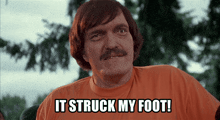 a man with a mustache wearing an orange shirt says " it struck my foot "