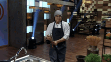 a chef wearing an apron and a beanie is holding a tong