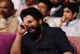 a man with a beard is sitting on a couch laughing while talking on a cell phone .