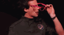 a man wearing a black shirt and pink sunglasses is sitting in front of a purple curtain .