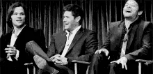 a black and white photo of three men in suits sitting next to each other on a stage .