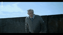 a man in a grey jacket and tie stands in front of a concrete wall with a blue sky behind him