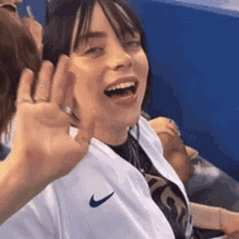 a woman wearing a white nike shirt is laughing and waving her hand .