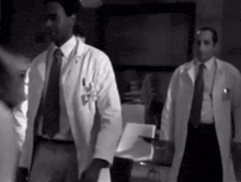 two men in lab coats and ties are walking in a dark room