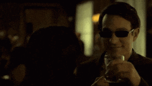 a man wearing sunglasses is drinking from a glass in a dark room .