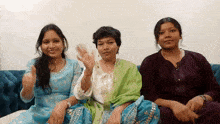 three women are sitting on a blue couch and one is waving
