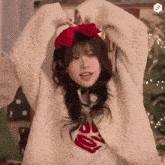 a girl wearing a white sweater and a red bow on her head stands in front of a christmas tree .