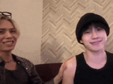 two men are sitting next to each other on a couch and smiling . one of the men is wearing a hat .