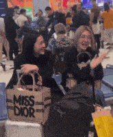 two women are sitting at an airport with a bag that says miss dior on it