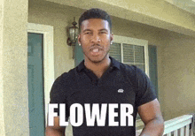 a man in a black shirt is standing in front of a house with the word flower written on his shirt .