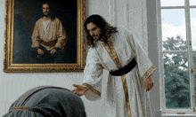 a man in a white robe with gold trim stands in front of a painting of jesus