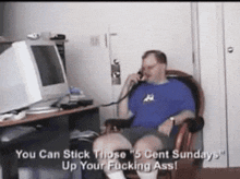a man in a blue shirt is sitting in front of a computer and talking on a cell phone