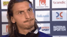 a man with long hair and a bow tie is standing in front of a wall with logos for scandic and folksam