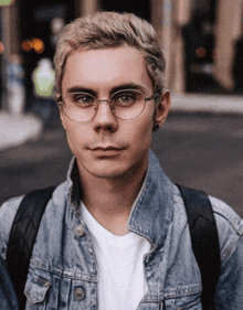 a young man wearing glasses and a denim jacket is looking at the camera