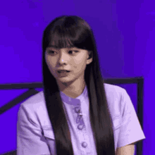 a woman with long hair is wearing a purple shirt and sitting in front of a blue background .