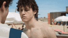 a shirtless man with curly hair is talking to another man on a beach
