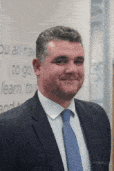a man in a suit and tie smiles in front of a wall that says " you all have to go learn to "