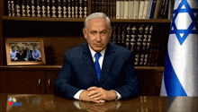 a man in a suit sits at a desk in front of a flag