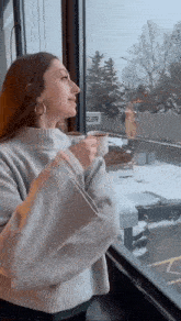 a woman in a white sweater is holding a cup of coffee and looking out a window .