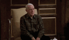 an elderly man in a plaid shirt is sitting in a chair with his arms outstretched .