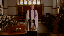 a priest stands in front of a coffin in a church with a stained glass window behind him