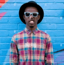 a man wearing a hat and sunglasses is sticking his tongue out in front of a colorful brick wall .