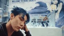 a man with blue hair is looking down in front of a welcome to hero society sign