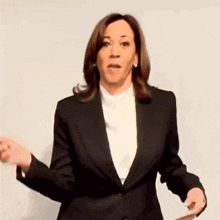 a woman in a black suit is standing in front of a white wall and making a funny face .