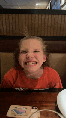 a young girl making a funny face while sitting at a table with a phone case that has a whale on it