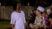 a man in a white shirt is standing next to another man in a white hat with the word man laksa on the bottom right