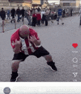 a bald man in a red shirt and black shorts is dancing on a sidewalk in front of a crowd .