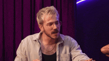 a man with blonde hair and glasses is sitting in front of a purple curtain and talking to someone .