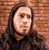 a man with long hair and a beard looks to the side in front of a brick wall