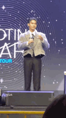 a man in a suit stands on a stage with a microphone in front of a sign that says ' tour '