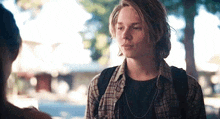 a young man wearing a plaid shirt and a necklace is talking to another young man .