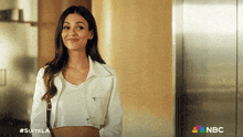 a woman in a white crop top is smiling in front of a nbc logo