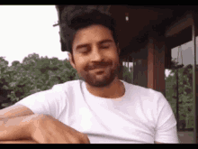 a man with a beard wearing a white t-shirt is smiling with his eyes closed