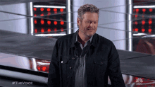 a man in a black jacket stands on a stage in front of a nbc logo