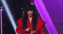 a woman in a red dress and black wig is standing on a stage with purple lights behind her .