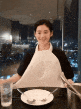 a woman wearing a white apron sits at a table with a plate and silverware