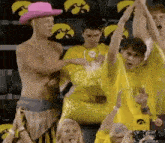 a shirtless man in a pink cowboy hat is standing in front of a crowd of people wearing yellow shirts .