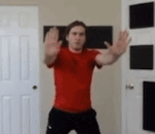 a man in a red shirt and black pants is standing in front of a door with his hands outstretched .