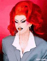 a drag queen with red hair is wearing a grey suit and white shirt