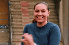 a woman in a blue sweater is smiling and clapping her hands in front of some stairs .