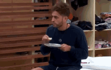 a man with a beard is sitting on a bed eating a plate of food .