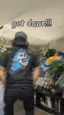 a man wearing a t-shirt with a skull on the back is standing in a living room and says get down