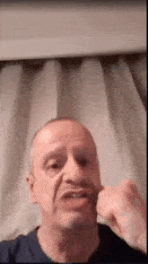 a man in a blue shirt is making a funny face with his fist in front of a white curtain .