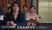 a woman is sitting at a desk in a classroom with other people and making a funny face .