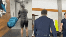 a man is carrying a blue bag and a man in a blue jacket is walking in a room .