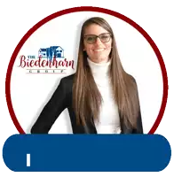 a woman wearing glasses and a turtleneck stands in front of a logo for the biedenharn group